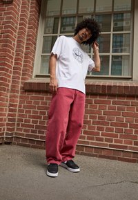 Witte T-shirt met korte mouwen en een grafisch ontwerp, gecombineerd met losse roze broek en zwarte schoenen, tegen een bakstenen muur.