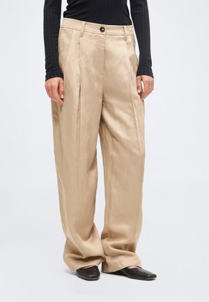 Pantalon beige à jambes larges avec plis devant, fermeture par bouton et fermeture éclair, porté avec un haut noir côtelé à manches longues et des chaussures plates noires.