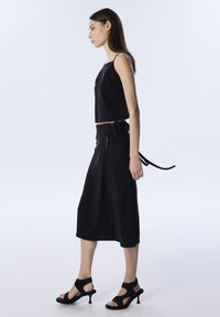 Top corto negro con tirantes finos, combinado con una falda negra de cintura alta que presenta un lazo en la cintura y detalles de botones, llevada con sandalias de tacón negras.