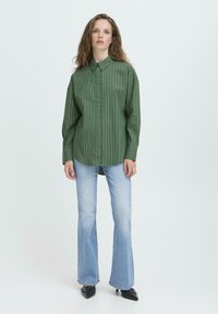 Žena s dlhými vlnitými vlasmi, oblečená v oversized zeleno-prúžkovanej košeli, svetlomodrých zvonových džínoch a čiernych špicatých topánkach, stojaca na jednokolorovom pozadí.