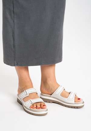 White slide sandals with two adjustable straps featuring round studs, a thick beige sole, and textured footbed. Red toenail polish visible.