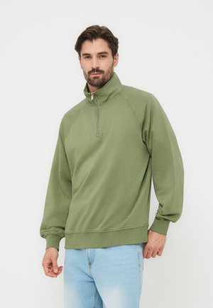 Mężczyzna w jasnozielonej bluzie z zamkiem do połowy i jasnoniebieskich dżinsach, stojący na jednolitym białym tle.