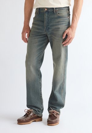 Hombre con vaqueros azules desgastados y zapatos marrones de cuero con cordones, de pie contra un fondo liso.