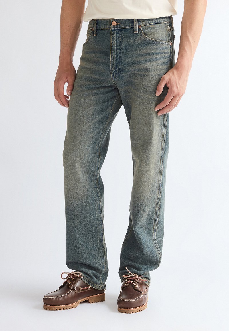 Homme portant un jean bleu délavé et des chaussures en cuir marron à lacets, debout devant un fond uni.