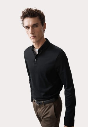 Camisa polo negra de manga larga con cuello y tapeta de tres botones, combinada con unos pantalones marrones y un cinturón negro.