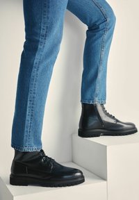 Czarne skórzane botki za kostkę z sznurowanym wzorem, teksturowane gumowe podeszwy i gładkie wykończenie, połączone z niebieskimi dżinsami na schodach.