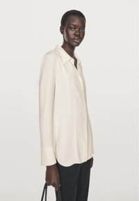 Langærmet creamfarvet bluse med knapper foran og krave. Har lige snit og blødt stof. Bruges med sorte bukser og en taske.