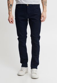 Sötétkék farmernadrág, slim fit fazonban, arany gombos záródással és négy zsebbel. Fehér sportcipővel viselve, sima textúrával.