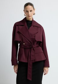 Bordeaux indpakningsjakke i en struktureret design med en stor krave, sidelommer og et bælte ved taljen, med en glat overflade.