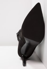 Black suede high-heeled shoe with a pointed toe, sleek design, and a textured black rubber sole with a narrow stiletto heel.