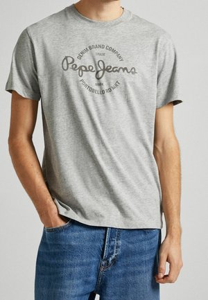 Mann trägt ein hellgraues, kurzärmeliges T-Shirt mit "Pepe Jeans"-Logo und Schriftzug, kombiniert mit blauen Jeans, eine Hand in der Tasche.