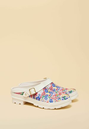 Pāris balti uzvelkami koka stila apavi ar daudzkrāsainiem ziedu rakstiem un regulējamām baltām siksnām ar zelta sprādzēm uz bēša fona.