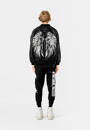 Chaqueta bomber negra con diseño de alas plateadas en la parte trasera, combinada con pantalones joggers negros que presentan gráficos de texto en blanco y acentos de salpicaduras. Botas blancas.