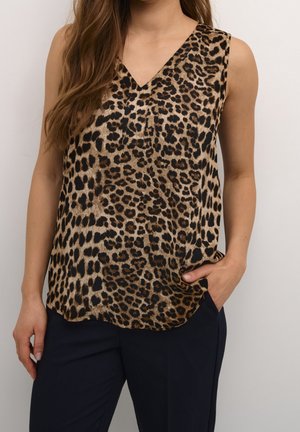 Vrouw draagt een mouwloze blouse met panterprint en V-hals, gecombineerd met een donkere broek, één hand in de zak, tegen een effen achtergrond.