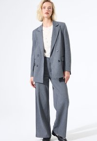 Traje gris a rayas con un blazer de doble botonadura y pantalones de pierna ancha, superpuesto a una camisa crema con pequeños adornos.