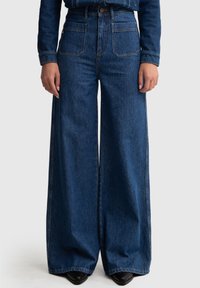 Pantalones de mezclilla de pierna ancha en azul oscuro, con dos bolsillos frontales, una cintura alta y costuras en contraste. El material tiene una textura suave.