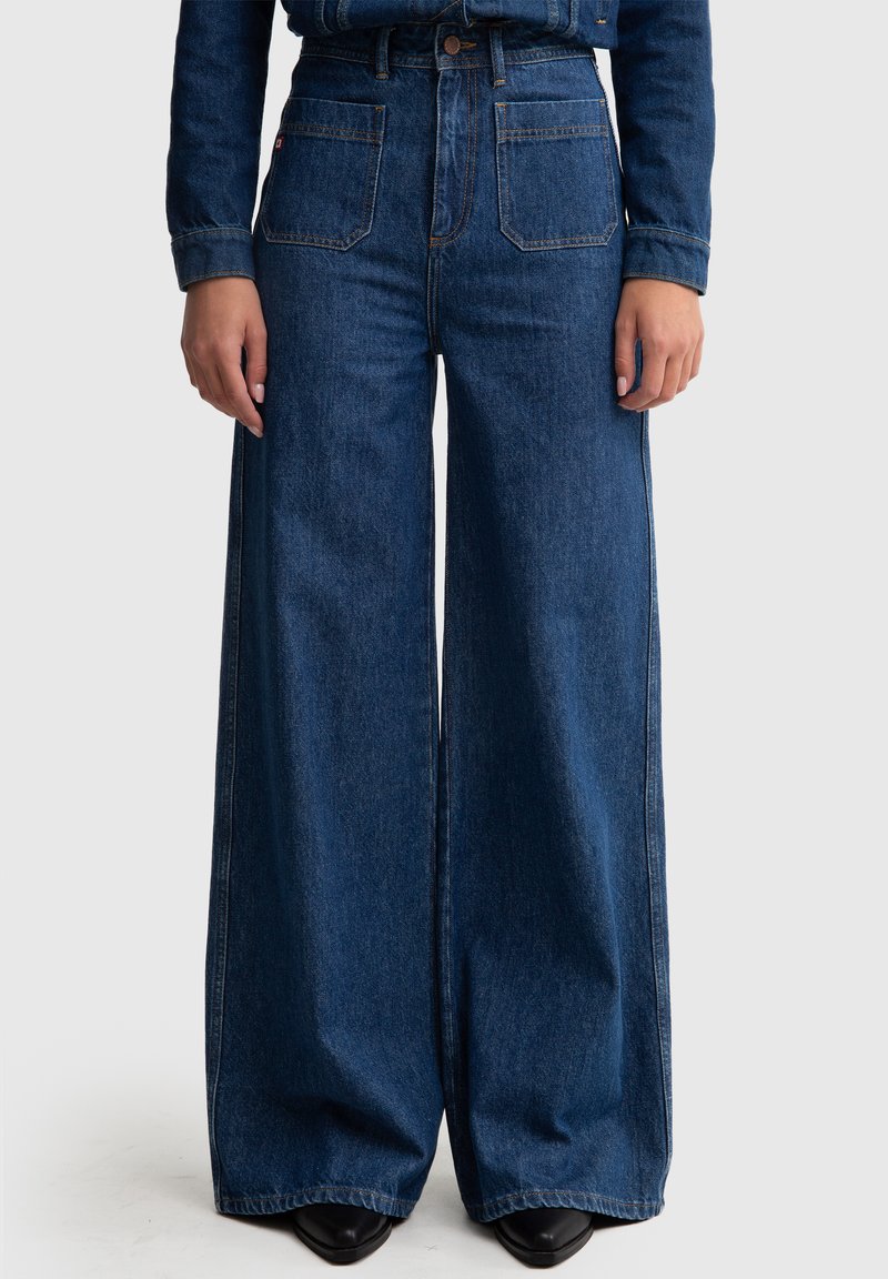 Pantalones de mezclilla de pierna ancha en azul oscuro, con dos bolsillos frontales, una cintura alta y costuras en contraste. El material tiene una textura suave.