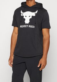 Musta lyhythihainen huppari, jossa on valkoinen härkälogo ja "PROJECT ROCK"-teksti. Valmistettu pehmeästä kankaasta, jossa säädettävät nyöritykset.