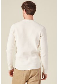 Weißer Strickpullover mit strukturiertem Muster, ausgestattet mit einem Rundhalsausschnitt und gerippten Bündchen, kombiniert mit hellbeigen Hosen.