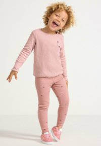 Vaaleanpunainen ribbattu pitkähihainen paita, jossa on pieni kukkakirjonta, yhdistettynä samanvärisiin vaaleanpunaiseen leggingsiin, joissa on kukka-applikaatioita. Valkoiset lenkkarit.