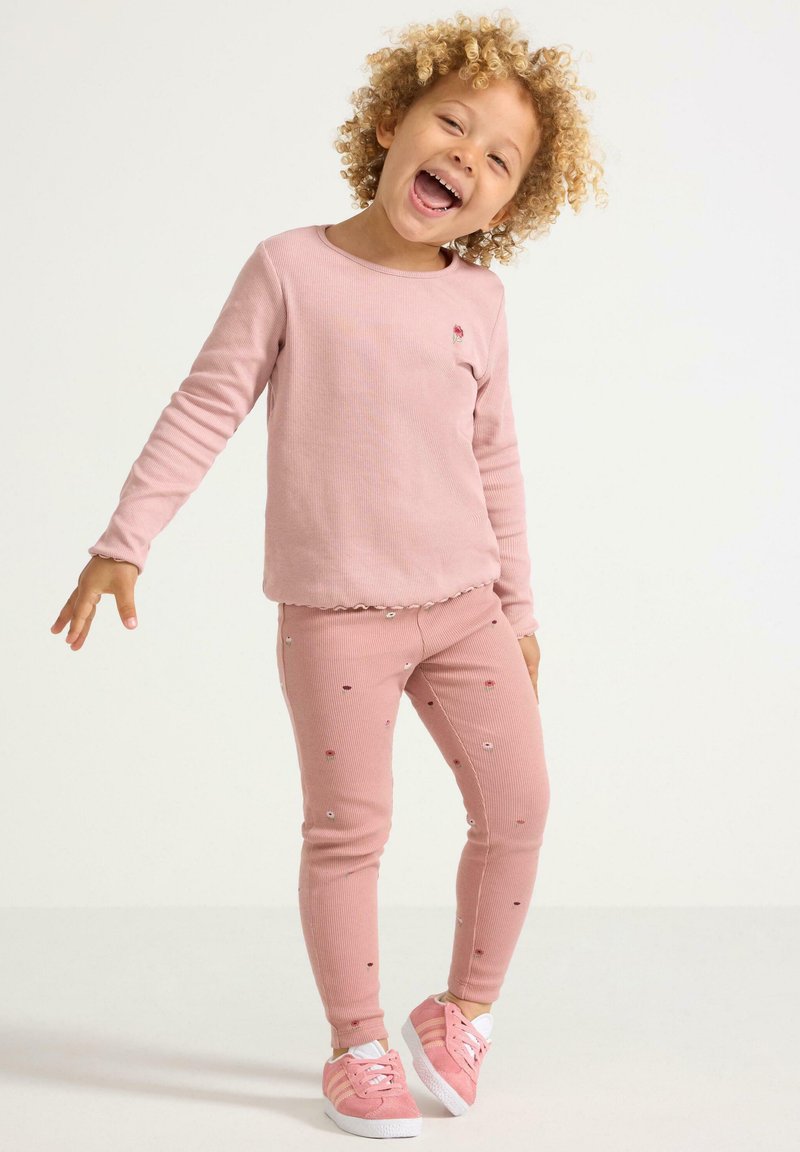 Vaaleanpunainen ribbattu pitkähihainen paita, jossa on pieni kukkakirjonta, yhdistettynä samanvärisiin vaaleanpunaiseen leggingsiin, joissa on kukka-applikaatioita. Valkoiset lenkkarit.