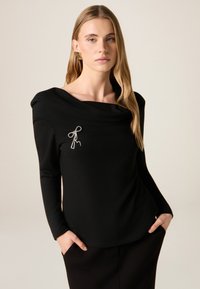 Zwart langärmelig shirt met een gedrapeerde halslijn en een zilveren strikdetail op de borst. De stof heeft een gladde textuur en biedt een ontspannen silhouet.