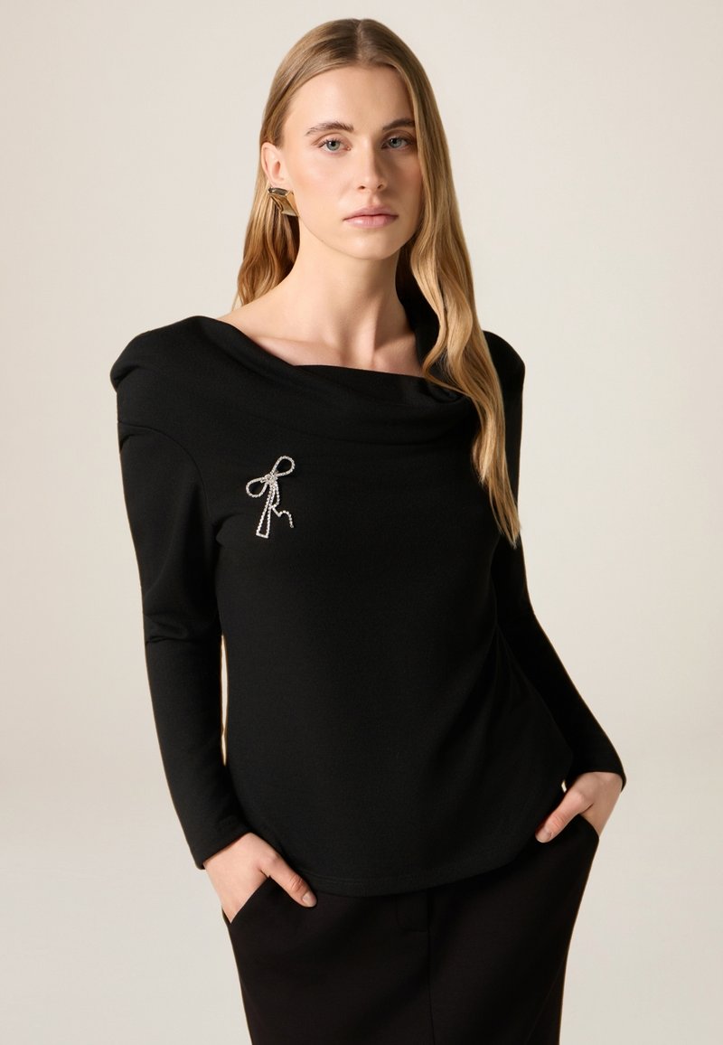 Zwart langärmelig shirt met een gedrapeerde halslijn en een zilveren strikdetail op de borst. De stof heeft een gladde textuur en biedt een ontspannen silhouet.