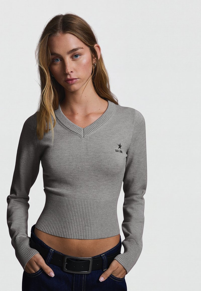 Femme aux longs cheveux châtain clair portant un pull gris ajusté à col en V et un pantalon sombre, les mains dans les poches, sur un fond clair uni.