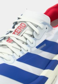 Tuva balta sporta apavu ar ziliem svītrām, baltiem šņorēm, sarkanu iekšpusi un uzrakstu "HATE ZERO" uz mēlītes.