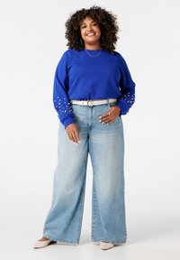 Blauwe sweatshirt met parelversieringen op de mouwen, gecombineerd met lichtblauwe wijde jeans en beige schoenen, op een witte achtergrond.