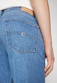 Šviesiai mėlyni džinsai su odiniu ženkleliu, ant kurio išspaustas žodis "JEANS", su viena užpakaline kišene, kurioje yra mažas logotipas ir matomi siuvimo apdailos elementai.