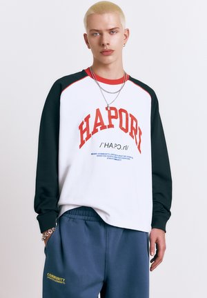 Ung person med kort blondt hår iført sort og hvid langærmet skjorte med rød tekst "HAPORI" og blå bukser med gul tekst.