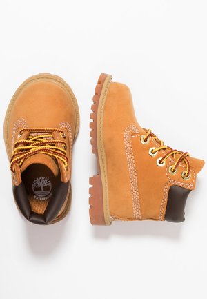 Paire de bottes en cuir imperméable couleur fauve avec semelles épaisses en caoutchouc, lacets jaunes et marron, et col rembourré noir sur fond blanc.