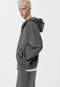 Sivá mikina s kapucňou a uvoľneným strihom, s rebrovanými manžetami a veľkým predným vreckom. Obsahuje zips a jemnú textúru.