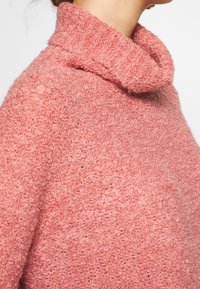 Pull rose doux avec col bénitier. Tissu texturé, poids moyen, coupe légèrement ample. Met en valeur un style décontracté.