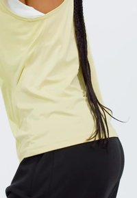 Camiseta deportiva de color amarillo claro con un ajuste holgado, que presenta un escote redondo y tela drapeada. Combinada con pantalones negros.