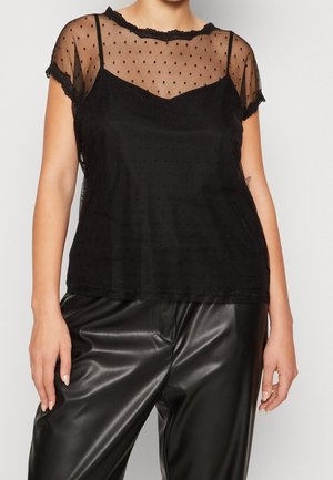 Femme portant un haut transparent à manches courtes noir à pois, par-dessus un débardeur noir, associé à un pantalon en cuir synthétique noir.