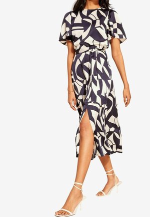 Woman wearing a black-and-white abstract patterned midi dress with short sleeves, side slit, waist tie, and white strappy heeled sandals.