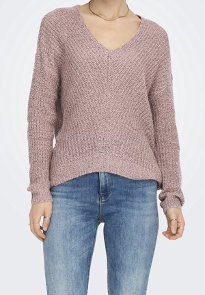 Mujer que lleva un suéter de punto color malva con cuello en V y mangas largas, y unos vaqueros azules de mezclilla, complementada con anillos y un collar fino de oro.