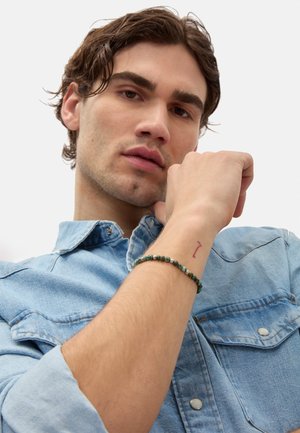 Beaded bracelet featuring green stones and silver accents worn on a wrist, close-up alongside a light blue denim shirt.