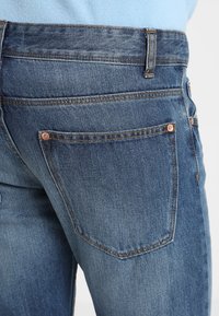 Närbild av en person som bär blå denimjeans som visar bakfickan, bältesöglan och kopparnitar, i kombination med en ljusblå skjorta.