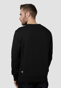 Felpa nera con collo rotondo, maniche lunghe e un sottile dettaglio del logo vicino all'orlo. Texture liscia, vestibilità casual e design semplice.