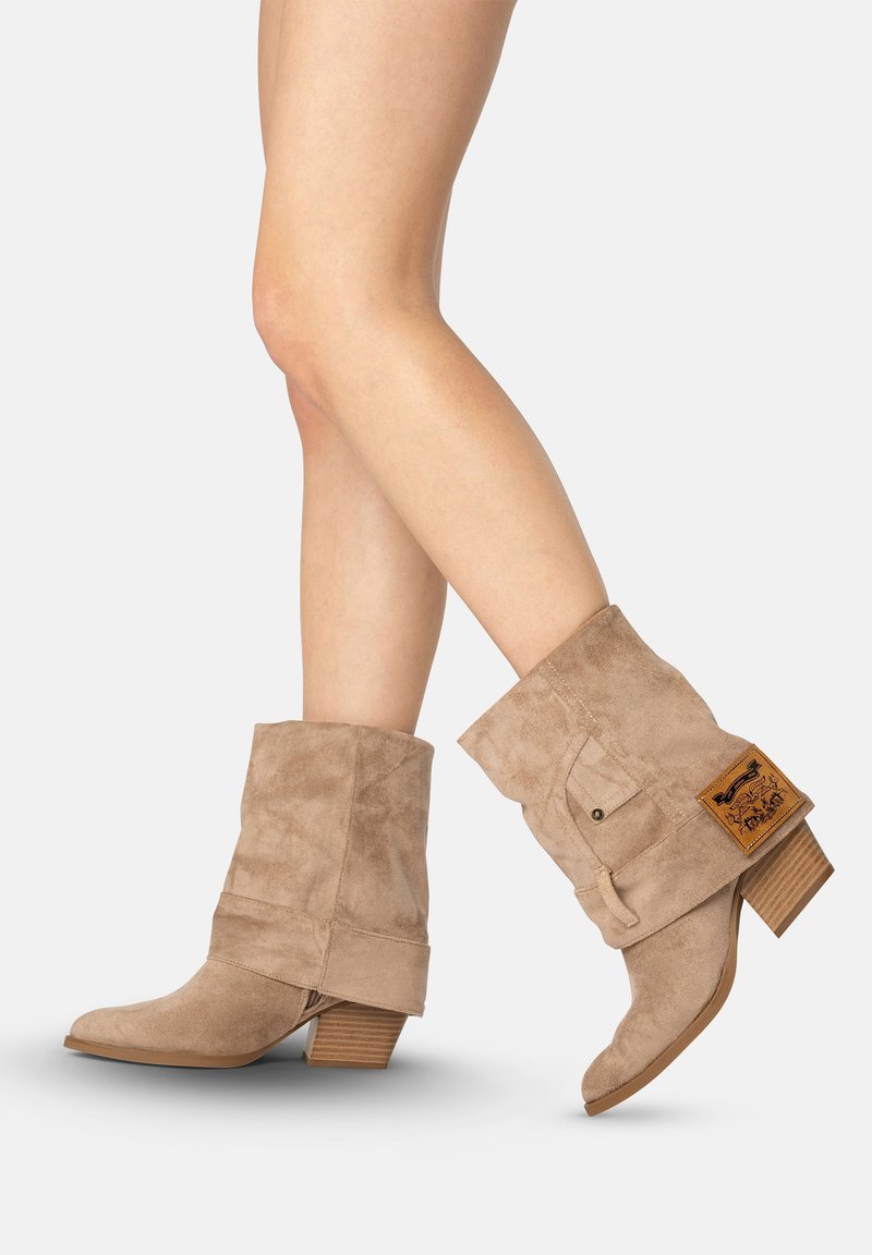 Piernas con botas marrón claro de ante hasta mitad de la pantorrilla, con tacones de bloque de madera y un parche de cuero en el lado exterior de la bota derecha.
