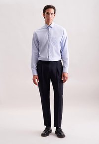 Camicia azzurra chiaro con bottoni e taschino, abbinata a pantaloni scuri a pieghe e scarpe nere. Tessuto liscio, vestibilità su misura.