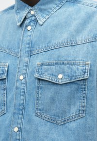 Ανοιχτό μπλε τζιν πουκάμισο με μύτη κολάρο, εμπρόσθια κλείσιμο με κουμπιά και δύο θησαυρούς στήθους με κλείσιμο σνάπ και ραμμένα λεπτομέρειες.