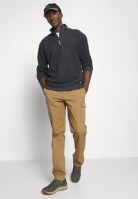 Grau gestrickter Pullover mit Reißverschlusskragen, kombiniert mit beige Cargo-Hosen mit Seitentaschen und grünen Wanderschuhen. Eine schwarze Mütze vervollständigt den Look.