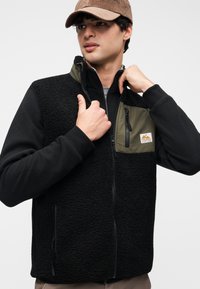 Μαύρο γιλέκο από φλις με πράσινες υφασμάτινες λεπτομέρειες, εμπρόσθια τσέπη με φερμουάρ, πλαϊνές τσέπες με φερμουάρ και υφή φινιρίσματος. Φορεμένο πάνω από γκρι μπλουζάκι.