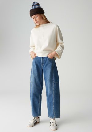 Cream-colored sweatshirt with wide sleeves, paired with high-waisted blue wide-leg jeans. Accented with a brown and blue striped knit beanie.