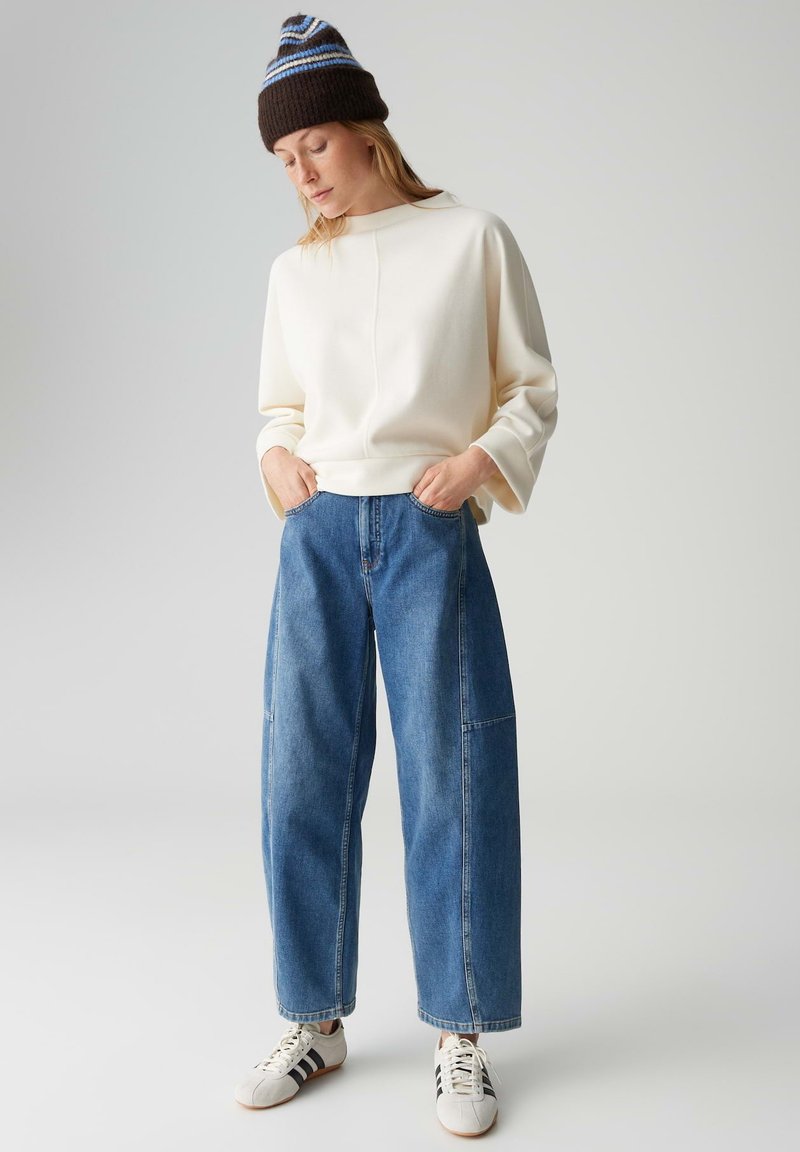 Cream-colored sweatshirt with wide sleeves, paired with high-waisted blue wide-leg jeans. Accented with a brown and blue striped knit beanie.