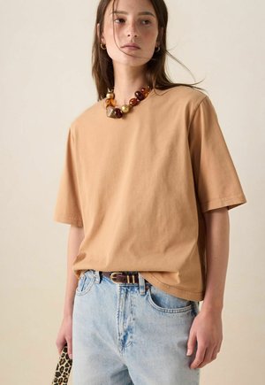 Femme portant une chemise beige à manches courtes, un jean bleu clair avec une ceinture foncée, un collier de perles ambrées, tenant un accessoire à imprimé léopard.